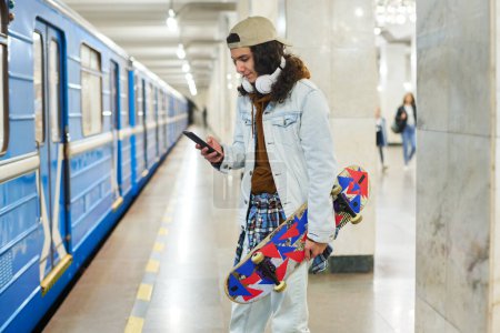 Photo for Teenage skateboarder in casual denim attire texting in mobile phone while standing in front of moving blue subway train before catching it - Royalty Free Image