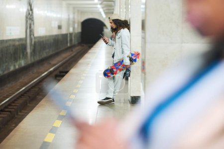 Photo for Focus on teenage guy with skateboard texting in smartphone while standing by marble column in front of railway at subway station - Royalty Free Image