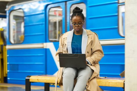 Foto de Joven mujer de negocios afroamericana con portátil sentado en el banco contra el tren azul del metro y la organización del trabajo o el uso de chat de vídeo - Imagen libre de derechos