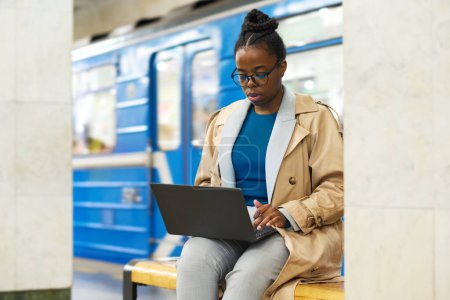 Foto de Joven mujer de negocios seria en casualwear creación de redes en la estación de metro mientras está sentado en el banco y mirando la pantalla del ordenador portátil contra el tren - Imagen libre de derechos