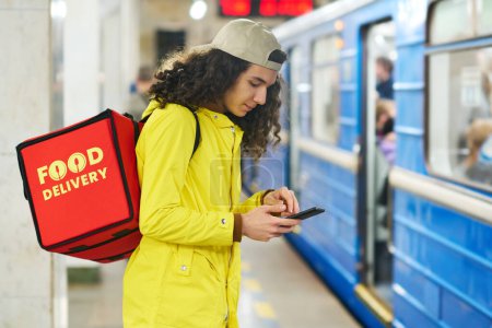 Foto de Joven repartidor en uniforme amarillo que envía mensajes de texto o se desplaza en el teléfono inteligente en la estación de metro mientras lleva una gran bolsa roja con comida en la espalda - Imagen libre de derechos