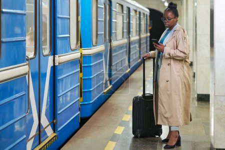 Foto de Joven mujer de negocios con la maleta de pie en la plataforma frente al tren azul en movimiento y mirando a través de datos en línea en el teléfono móvil - Imagen libre de derechos