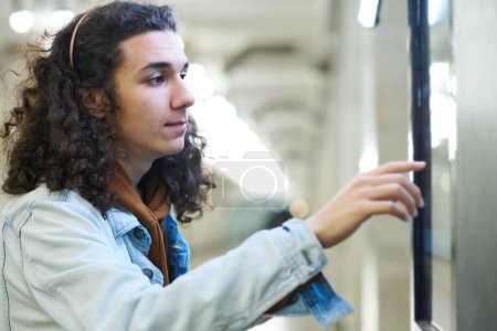 Photo for Side view of teenager in denim jacket pointing at interactive display with timetable or route map of trains while standing at subway station - Royalty Free Image