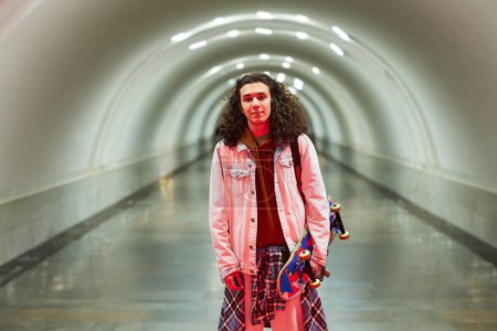 Foto de Elegante joven con monopatín en la mano de pie delante de la cámara contra un largo túnel de la estación de metro o metro y mirándote - Imagen libre de derechos