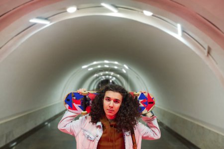 Photo for Youthful guy holding skateboard behind his head and looking at camera while standing on background of long tunnel in subway station - Royalty Free Image