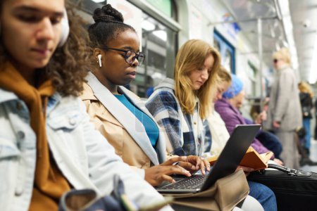 Foto de Joven mujer de negocios seria en auriculares escribiendo en el teclado del ordenador portátil mientras está sentado entre otros pasajeros que viajan en tren subterráneo - Imagen libre de derechos