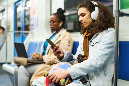 Foto de Adolescente serio en auriculares mensajes de texto en el teléfono móvil mientras está sentado en el tren subterráneo contra la mujer de negocios afroamericana con computadora portátil - Imagen libre de derechos