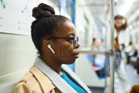 Téléchargez les photos : Vue latérale de la jeune femme afro-américaine sérieuse en lunettes et vêtements décontractés écouter de la musique dans les écouteurs tout en voyageant en métro - en image libre de droit
