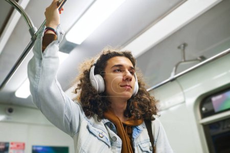 Photo for Smiling teenager in denim jacket holding by handrail in subway train while riding to school in the morning and listening to music in headphones - Royalty Free Image