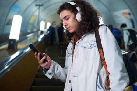 Photo for Teenage guy in casualwear and headphones using smartphone while standing on escalator, watching video and moving downwards - Royalty Free Image