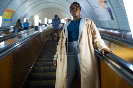 Foto de Joven mujer afroamericana en ropa de casualwear de pie sobre escaleras mecánicas y moviéndose hacia abajo contra el largo túnel de metro con otras personas - Imagen libre de derechos