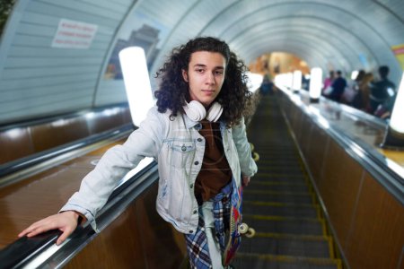 Photo for Serious teenage guy with skateboard looking at camera while standing on moving escalator and riding downwards to subway station - Royalty Free Image