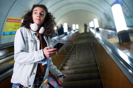 Photo for Teenage skateboarder with smartphone standing on moving escalator and looking aside against long tunnel and interior of underground - Royalty Free Image