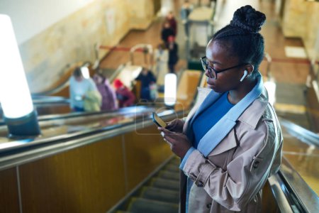 Foto de Vista lateral de la joven mujer negra en gafas viendo vídeo en línea en el teléfono inteligente mientras se mueve hacia abajo en escaleras mecánicas en el metro - Imagen libre de derechos