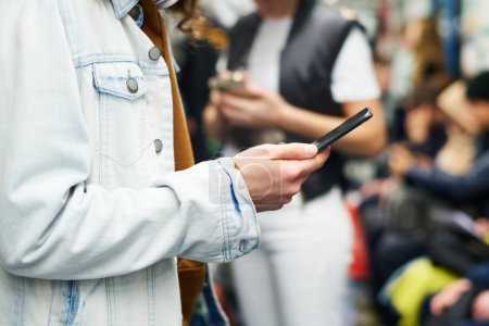 Photo for Close-up of young man in denim jacket texting in mobile phone or watching video while standing in front of camera against crowd of people - Royalty Free Image