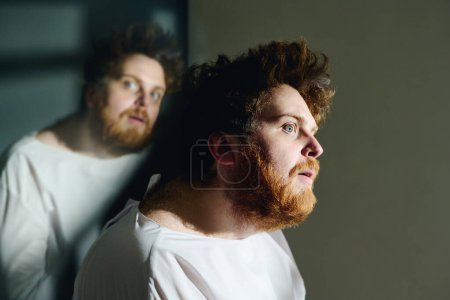 Photo for Side view of young insane man in white straitshirt sitting in front of camera against his reflection and looking forwards with anxious expression - Royalty Free Image