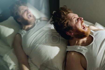 Foto de Doble exposición de un joven loco en chaleco gritando en voz alta de horror o durante la agonía mientras está acostado en la cama en la sala de psiquiatría - Imagen libre de derechos