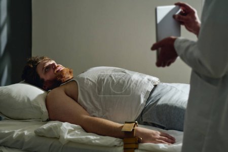 Photo for Young male patient of mental hospital lying in bed with his wrists bound with tight belts and looking at screen of tablet held by doctor - Royalty Free Image