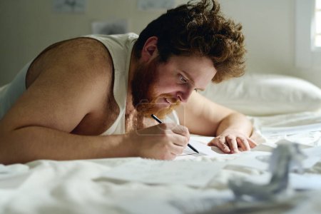 Téléchargez les photos : Vue latérale du jeune homme barbu en gilet se penchant sur le lit tout en dessinant quelque chose sur papier avec crayon au cours du traitement mental - en image libre de droit
