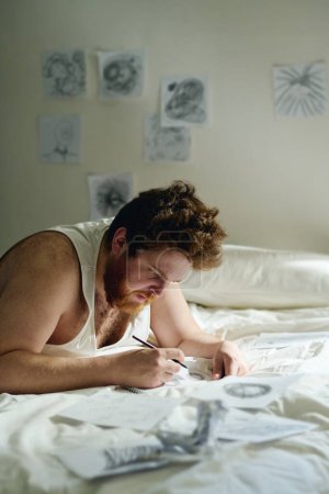 Photo for Vertical shot of bearded male patient of mental hospital drawing with pencil on paper sheets while bending over bed in clinics ward - Royalty Free Image