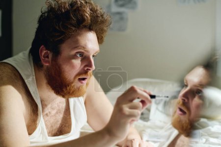 Photo for Young fat bearded man with depression or narcissistic mania bending ober bed in front of camera and pointing at reflection of his face - Royalty Free Image