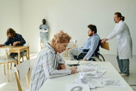 Photo for Side view of mature blond female patient of lunatic asylum drawing something on paper sheet while sitting by desk against other people - Royalty Free Image