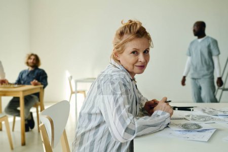 Photo for Mature insane woman in pajamas looking at camera while sitting by desk against male patient and doctor of mental hospital and drawing - Royalty Free Image