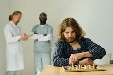 Photo for Bearded male patient of mental hospital in pajamas playing chess on his own while sitting by desk against two intercultural psychiatrists - Royalty Free Image