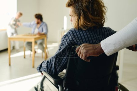 Photo for Rear view of patient of mental hospital with anxiety disorder sitting in wheelchair while African American caregiver pushing him forwards - Royalty Free Image