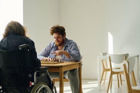Photo for Young bearded man in pajamas pointing at pawn while playing chess with other patient of mental hospital sitting in wheelchair in front of him - Royalty Free Image