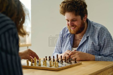 Foto de Joven paciente masculino sonriente de clínicas mentales mirando al tablero de ajedrez mientras su compañero de juego hace movimiento y pone peón blanco en otro cuadrado - Imagen libre de derechos