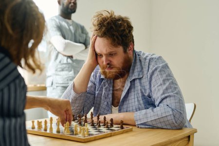 Foto de Hombre nervioso con peón en la nariz mirando al compañero de ajedrez haciendo movimiento mientras ambos están sentados uno frente al otro por una mesa de madera - Imagen libre de derechos