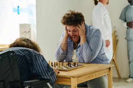 Photo for Nervous male patient of mental hospital touching head and screaming during game of chess while looking at pawns on chessboard - Royalty Free Image