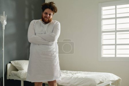 Téléchargez les photos : Jeune homme en surpoids en camisole de force blanche debout devant la caméra contre son lit dans la salle de l'hôpital psychiatrique le soutenant - en image libre de droit
