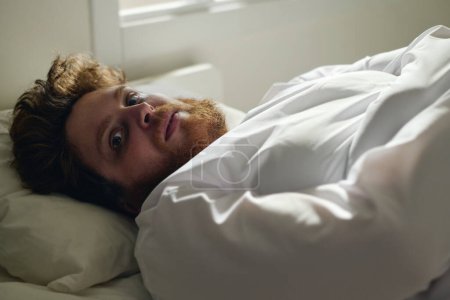 Téléchargez les photos : Jeune fou patient masculin de cliniques mentales regardant la caméra avec expression faciale dérangée tout en étant couché dans le lit pendant la procédure médicale - en image libre de droit