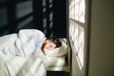 Foto de Joven hombre trastornado con camisa de fuerza mordiendo almohada mientras está acostado en la cama por ventana en el hospital psiquiátrico en aislamiento durante la terapia - Imagen libre de derechos