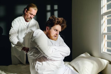 Mature psychiatrist standing by bed of insane male patient suffering from psychosis and tying sleeves of straitjacket on his back in mental hospital
