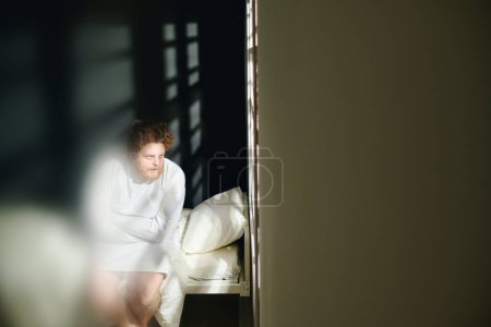 Photo for Focus on young deranged man suffering from strong mental disability sitting on bed in isolation in hospital ward and looking through window - Royalty Free Image