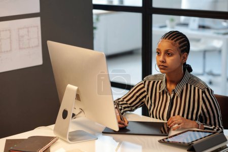 Téléchargez les photos : Jeune webdesigner afro-américain ou artiste avec stylet dessin croquis sur écran tactile tout en étant assis par le lieu de travail devant l'ordinateur - en image libre de droit