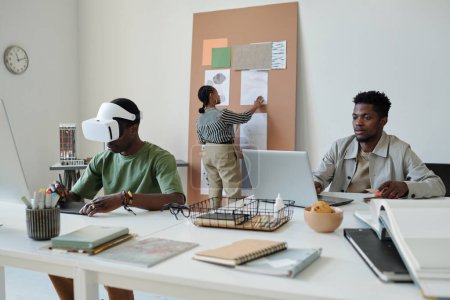 Téléchargez les photos : Jeune webdesigner afro-américain regardant l'écran d'ordinateur portable tout en étant assis par le lieu de travail à côté de son collègue dans le casque créant de nouveaux jeux informatiques - en image libre de droit