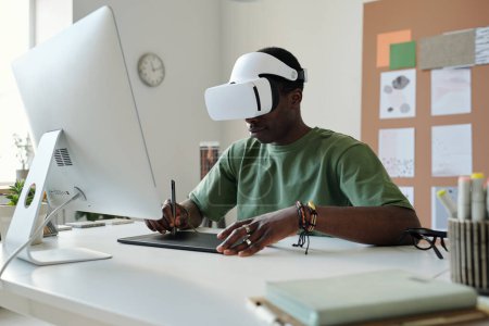 Téléchargez les photos : Jeune designer masculin en réalité virtuelle dessin de casque croquis graphiques sur le pavé tactile avec stylet tout en étant assis devant l'écran d'ordinateur - en image libre de droit