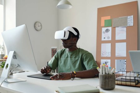 Junge Grafikdesignerin im erweiterten Reality-Headset mit Stift und Touchpad bei der Erstellung neuer Computerspiele am Arbeitsplatz