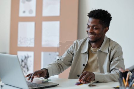 Foto de Joven sonriente afroamericano diseñador web masculino presionando la tecla del teclado del ordenador portátil mientras está sentado en el lugar de trabajo y la creación de un nuevo sitio web - Imagen libre de derechos