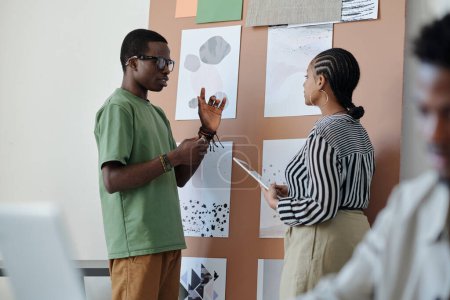 Foto de Diseñador o arquitecto masculino de confianza explicando algo a una colega por tablón de anuncios con un grupo de papeles con bocetos gráficos - Imagen libre de derechos