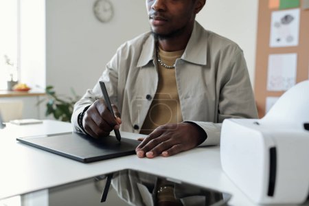 Photo for Cropped shot of young African American male designer drawing graphics while sitting by workplace and holding stylus on touchpad screen - Royalty Free Image