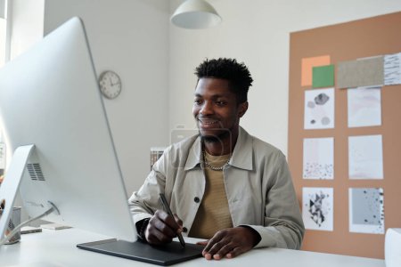 Téléchargez les photos : Jeune graphiste afro-américain souriant ou retoucher avec stylet et pavé tactile regardant l'écran d'ordinateur pendant le travail au bureau - en image libre de droit