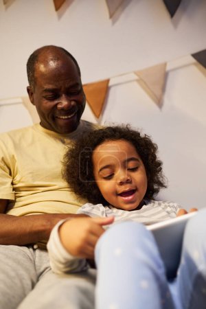 Téléchargez les photos : Image verticale du père afro-américain utilisant la tablette pc avec son fils au lit - en image libre de droit