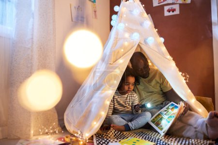 Téléchargez les photos : Papa lisant le livre à son enfant alors qu'ils sont assis dans une tente faite à la main dans la chambre - en image libre de droit