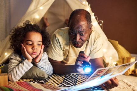 Téléchargez les photos : Afro-américain papa lecture livre à son petit fils en utilisant une lampe de poche alors qu'ils sont couchés sur le sol dans la tente maison - en image libre de droit