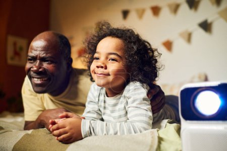 Téléchargez les photos : Joyeux papa regardant un film sur projecteur avec son petit fils avant de dormir dans la chambre - en image libre de droit